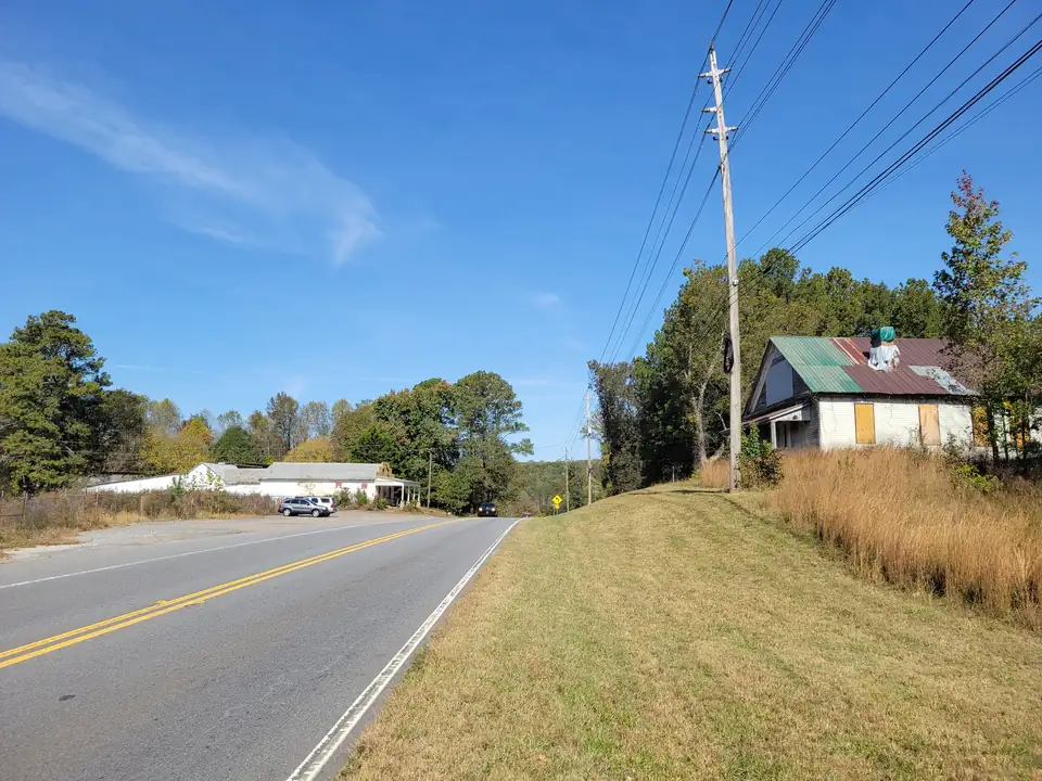 Two-lane highway curving past a grassy bank and the old white homestead, with an old white store building in the distance, autumn trees and power lines