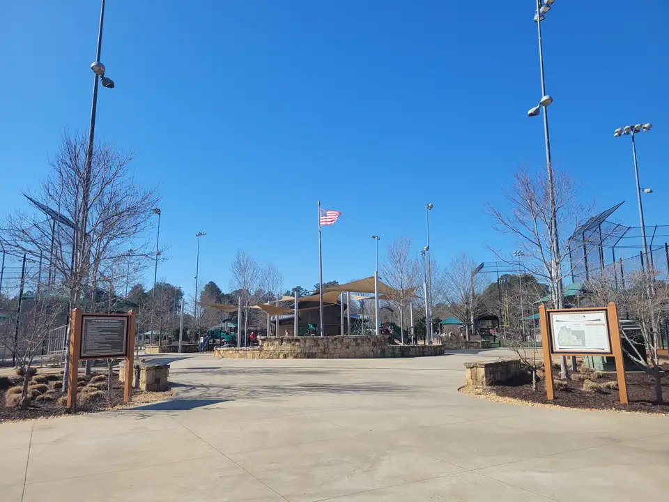 Wide concrete entry walk between wooden info kiosks leading to shaded ballfield seating, light towers and an American flag, bare early-spring trees, clear blue sky