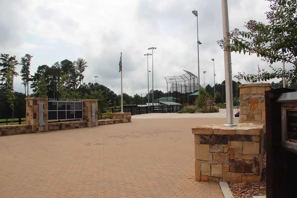 Brick-paver plaza with stone pillars and a dark memorial wall, flagpoles and ballfield light towers and backstops beyond, cloudy sky