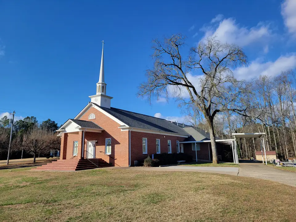 Red-brick church with a white steeple and portico on a broad lawn, a huge bare oak beside it and a covered walkway toward the cemetery, blue sky with clouds