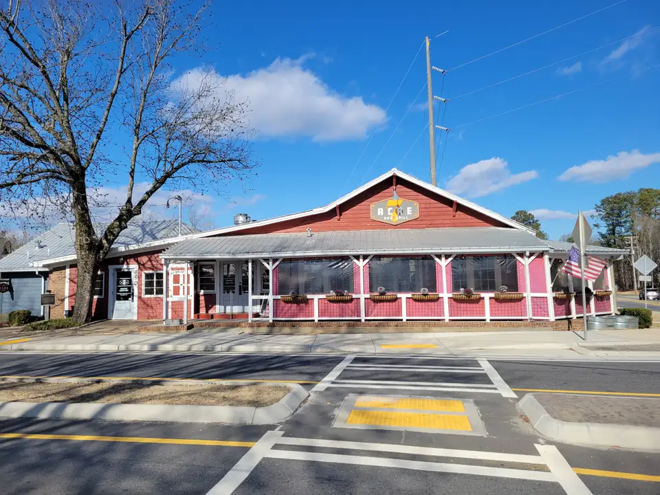 Red wooden building with a white-trimmed screened porch, window boxes, a '7 Acre Bar & Grill' sign and an American flag, a historic marker by the side door, new crosswalk in front, bare tree and blue sky