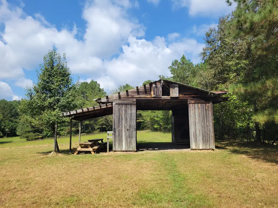 Weathered gray board barn with open bays and a lean-to sheltering picnic tables, set in a mown meadow edged by pines, puffy white clouds