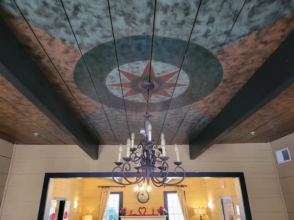 Interior: a wooden plank ceiling painted with a red eight-point star in green and cream rings over sponge-painted panels, an iron chandelier, a doorway into a yellow room with a mantel