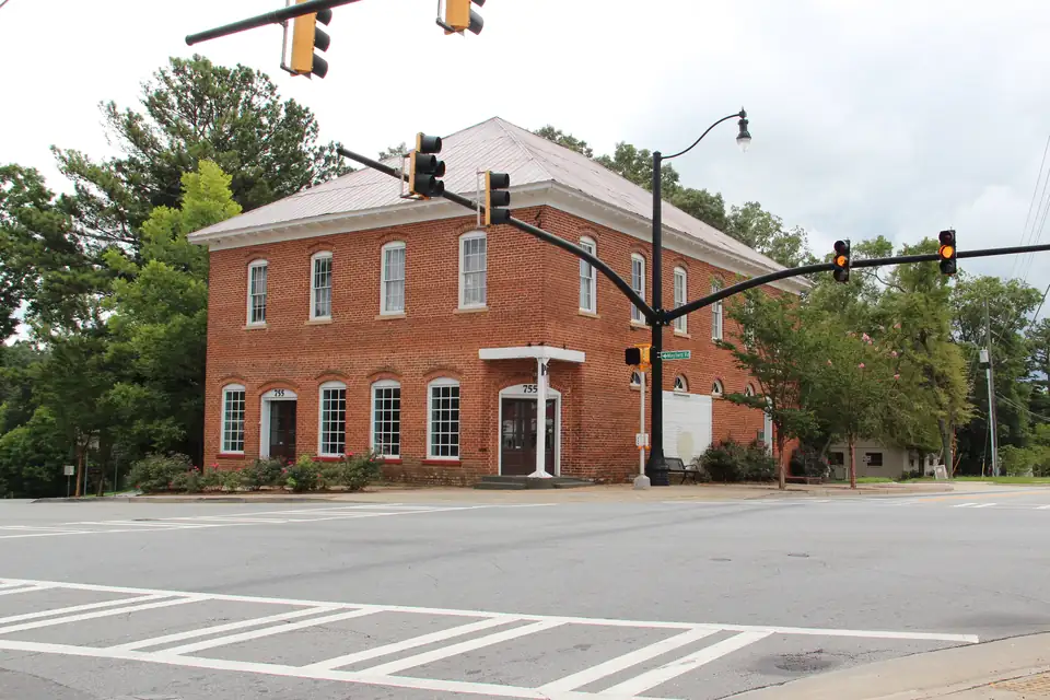 Two-story red-brick building with arched windows, hipped metal roof and two doors numbered 755, seen across a wide intersection with traffic signals and a 'Mayfield Rd' street sign; overcast summer sky