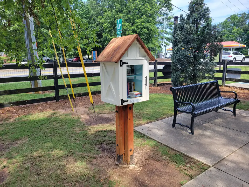 White barn-style Little Free Library box with a cedar roof on a wooden post, a black metal bench, black board fence and a road beyond; a gas-station canopy is visible in the far background