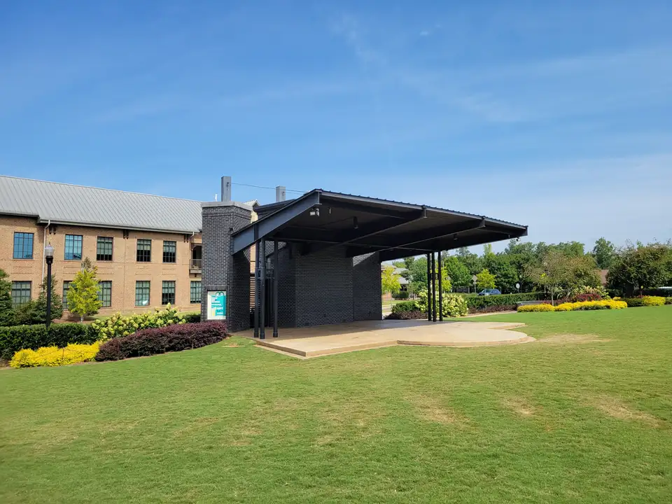 Covered black-brick outdoor stage facing a large green lawn, with the brick City Hall building to the left and flowering shrubs; a 'Shop Sip & Stroll on the Green' event poster on the stage pier