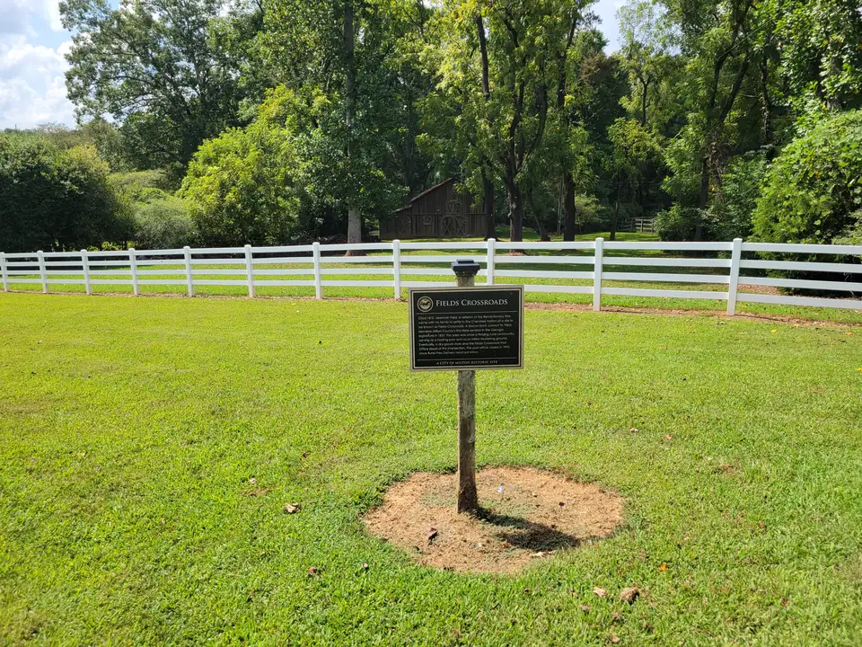 Bronze-on-black 'FIELDS CROSSROADS — A City of Milton Historic Site' marker on a post in a green lawn, a long white four-rail fence and a weathered wooden barn under big trees