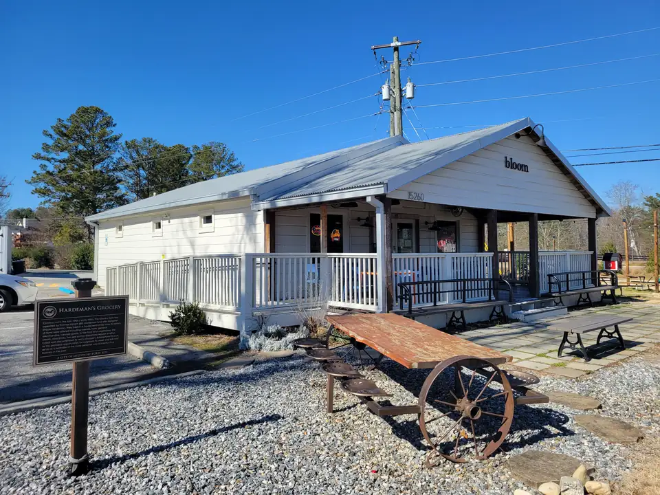 Restored white wooden store with a metal roof and wraparound porch, 'bloom' sign and number 15260, a 'Hardeman's Grocery' historic marker and a wagon-wheel picnic table on gravel, clear winter sky