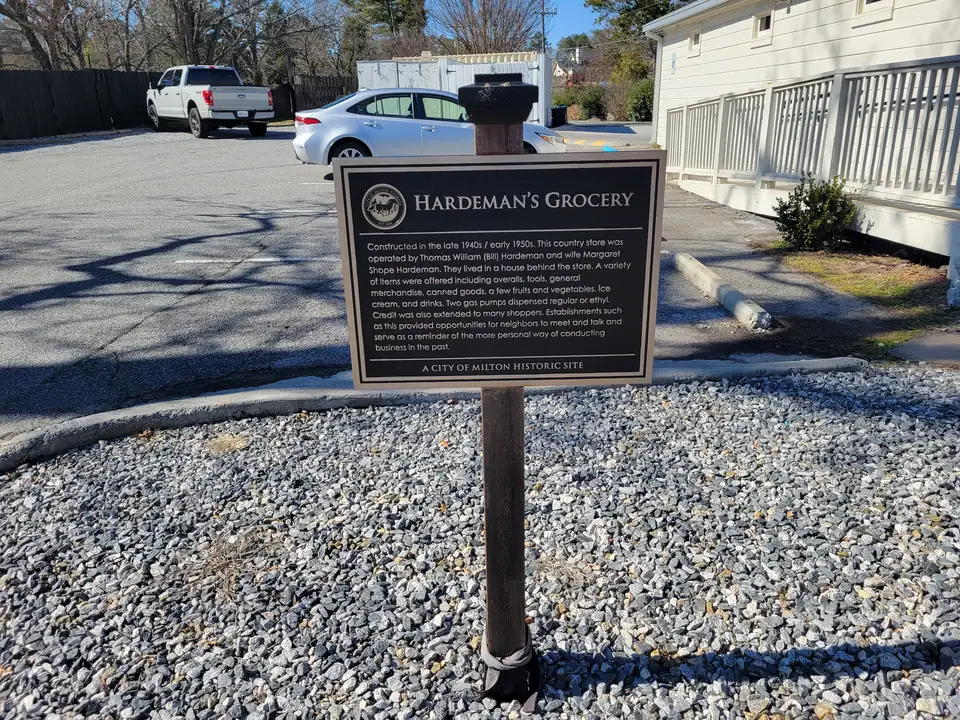 Black 'Hardeman's Grocery — A City of Milton Historic Site' plaque on a post in gravel, parking lot with cars and the white store porch behind
