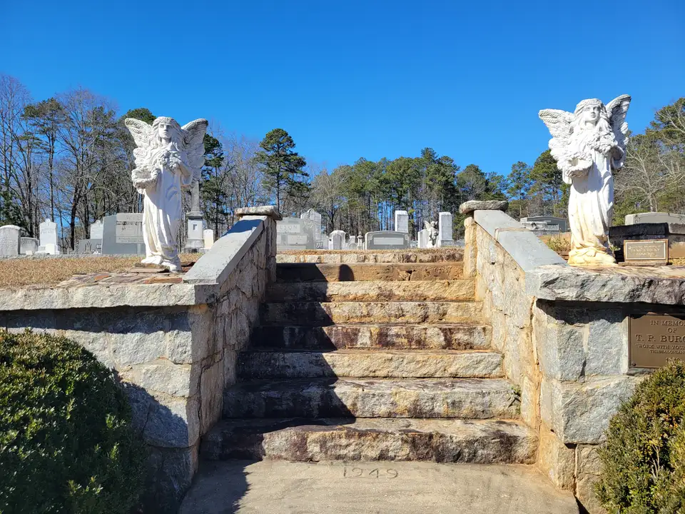 Granite steps flanked by fieldstone walls and two white angel statues rising to rows of headstones, bare winter trees and pines, '1949' carved in the bottom step; memorial plaques with family names
