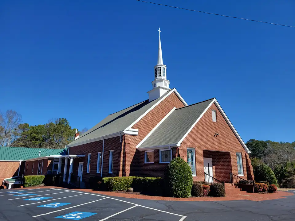 Red-brick church with a tall white steeple and cross, stained-glass windows, clipped shrubs and a paved lot with accessible spaces under deep blue winter sky