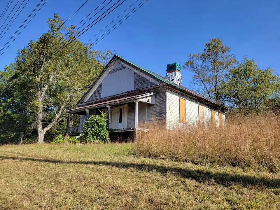 Weathered white clapboard farmhouse with a front porch, boarded windows and a green metal roof, tall golden grass and trees, power lines overhead, blue sky
