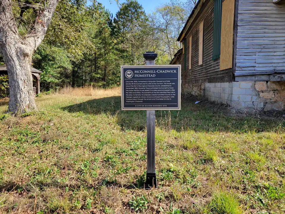 Black City of Milton historic-site plaque on a wooden post in grass; text about the 1832 land lottery, the first merchant flour mill and the proposal of Milton County