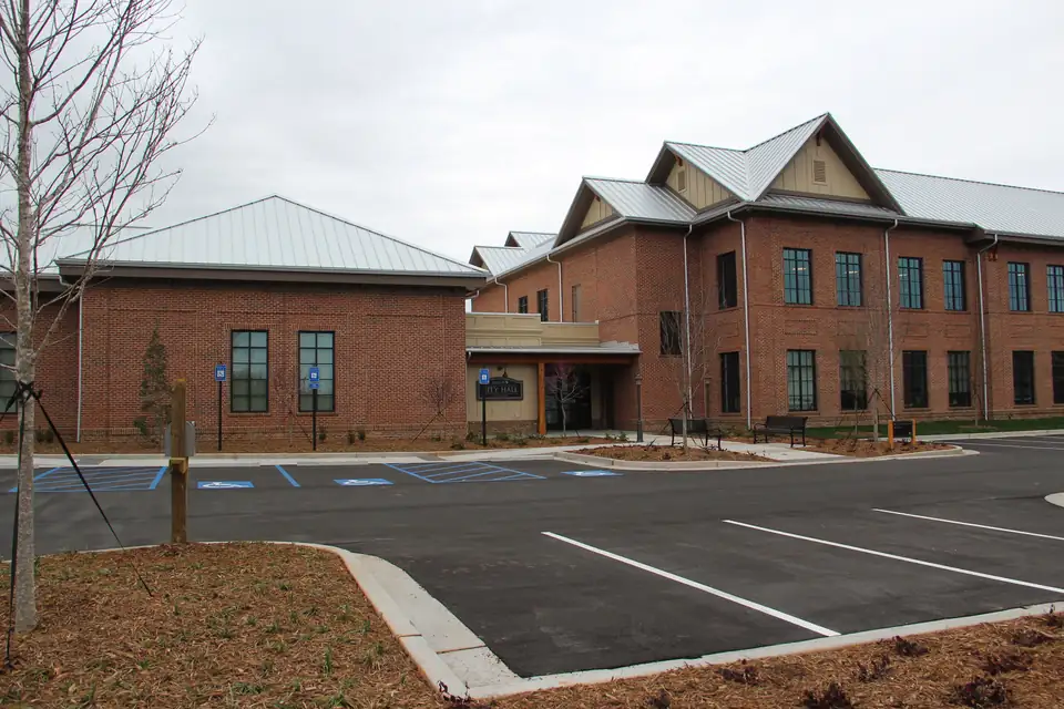 Brick city hall with standing-seam metal roofs and a 'CITY HALL' sign at the entrance, fresh parking lot and young trees under a gray sky