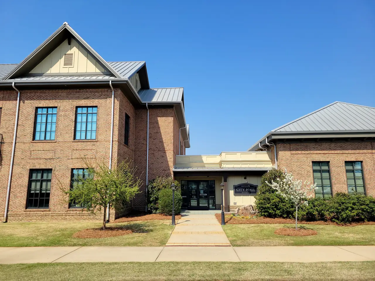 Front walk leading to the glass entrance of the brick city hall, 'MILTON CITY HALL' sign, a white flowering tree and a lawn under clear blue sky