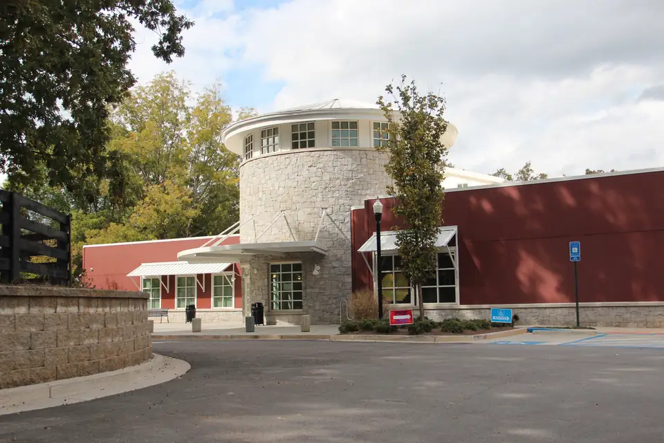 Round stone entrance tower with a white cap and clerestory windows between red board-and-batten wings; black horse-country fence at left; autumn-tinged trees; a 'VOTE HERE' sign by the door