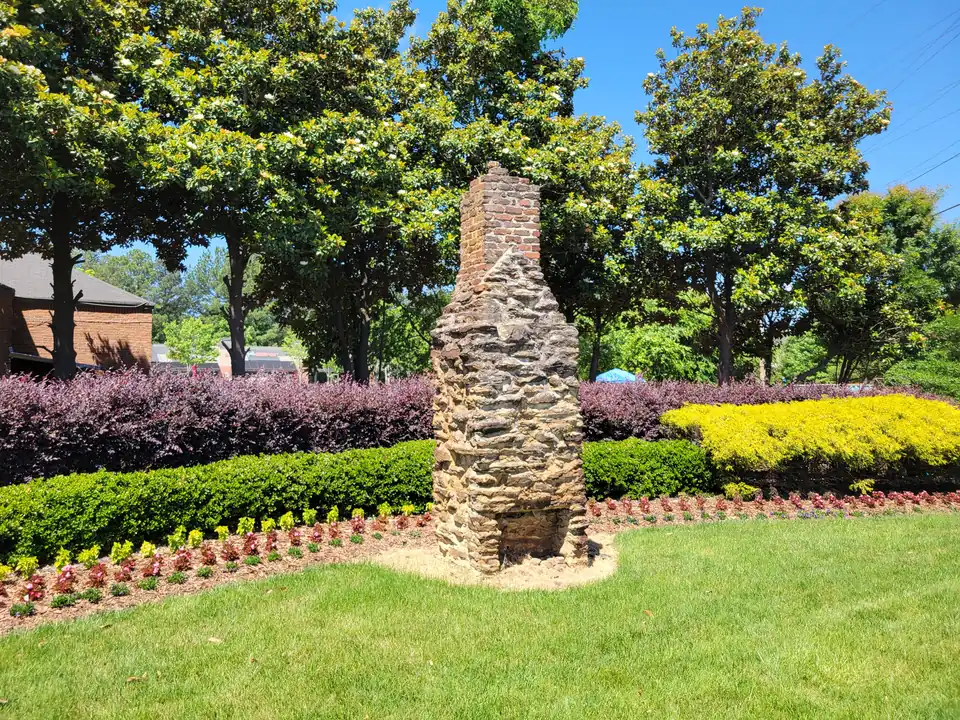 Freestanding rubble-stone and brick chimney standing in a mulched bed of red and yellow plantings and purple shrubs, magnolias behind, blue sky