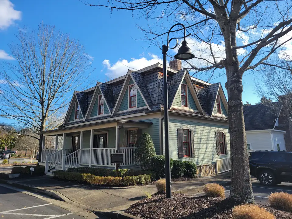 Sage-green two-story house with steep shingled gables, red window sashes, a white porch and a 'Thomas B. Newton House' historic marker, lamppost and bare trees; a parked SUV at right; a small home-security yard sign