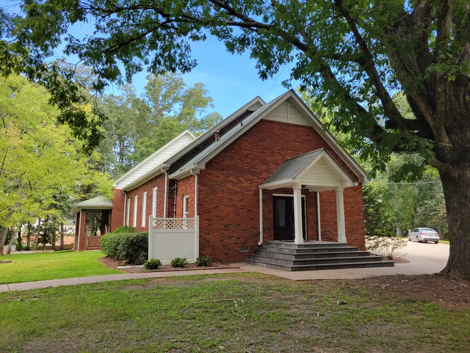 Red-brick gable-front church with a white columned porch and steps, shaded by a large oak, gravel drive and one parked car