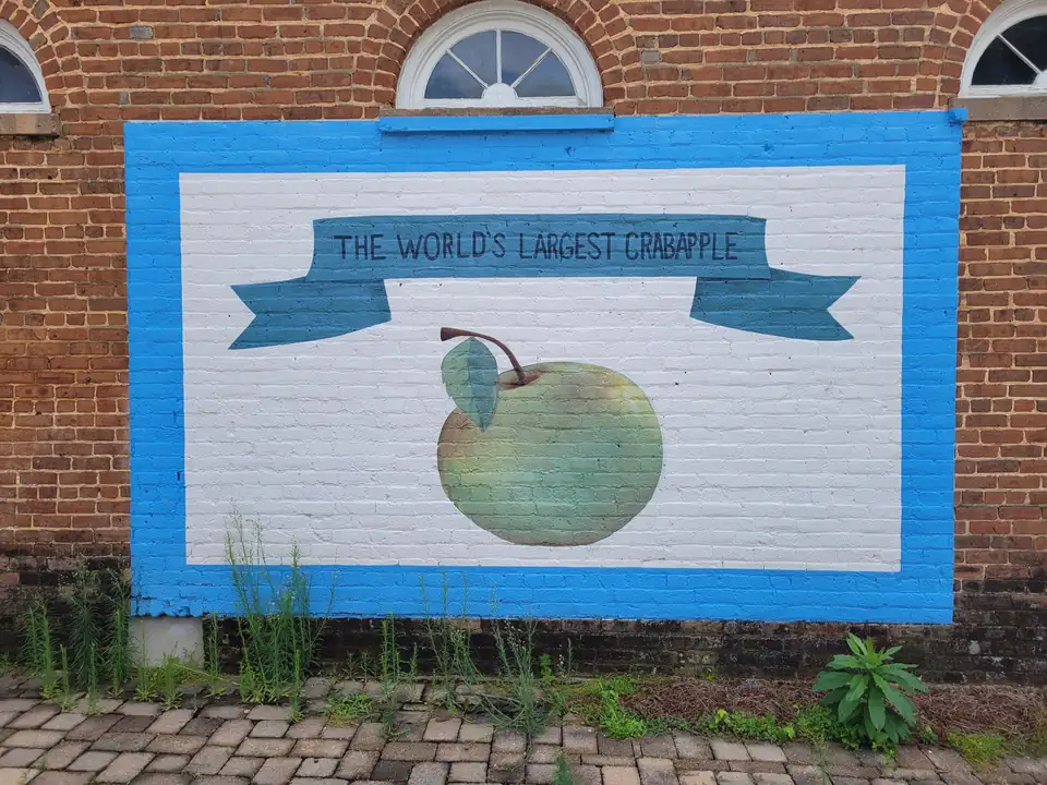 Painted sign on old red brick: a green crabapple under a blue ribbon reading 'THE WORLD'S LARGEST CRABAPPLE', framed in bright blue, arched fanlight windows above, weeds along a brick-paver walk